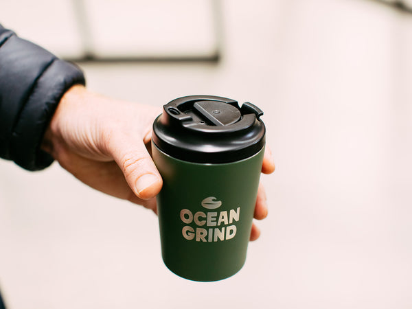 Hand holding a Khaki 'Ocean Grind' coffee cup with a black click lid.
