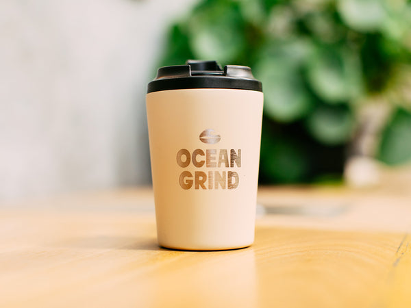 Oat Fressko cup with 'Ocean Grind' logo on a blurred natural background
