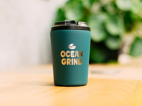 Emerald coffee cup with 'Ocean Grind' logo on a blurred natural background