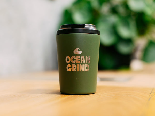 Kahki coffee cup with 'Ocean Grind' logo on a blurred natural background