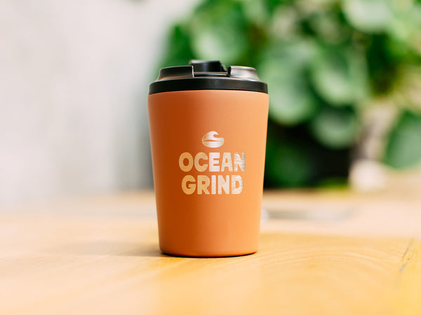 Tuscan Fressko cup with 'Ocean Grind' logo on a blurred natural background
